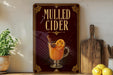 A framed advertisement for "Mulled Cider" featuring an illustration of a glass mug filled with a warm, spiced beverage garnished with a slice of lemon and a cinnamon stick.