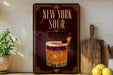 A decorative sign for "New York Sour" cocktail, featuring a glass illustration and a cherry on top, placed on a wooden surface with a potted plant and a cutting board nearby.