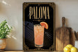 A framed advertisement for a cocktail called "Paloma" featuring a glass of the drink with a slice of orange on the rim.