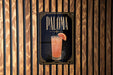 A framed poster of a cocktail called "Paloma" is displayed on a wooden wall.