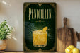 A framed advertisement for a drink called "Penicillin" with a glass of the drink and lemon slices, placed on a wooden shelf next to a cutting board and a potted plant.