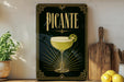 A framed poster of a cocktail with the word "PICANTE" prominently displayed, along with a decorative border and a glass illustration. The poster is placed on a shelf next to a wooden cutting board and a potted plant.
