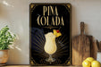 A decorative sign for a Pina Colada cocktail, featuring a glass of the drink and a pineapple garnish, is displayed on a wooden shelf next to a potted plant and a cutting board.