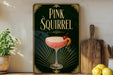 A framed poster of a pink cocktail with a cherry on top, displayed on a wall next to a wooden cutting board and a potted plant.