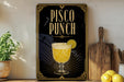 A framed poster of a pisco punch drink is displayed on a wall, with a wooden cutting board and a potted plant to the side.