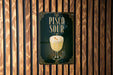 A poster with the text "Pisco Sour" and an illustration of a glass of pisco sour on a wooden wall.