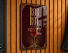 A sign for "Rob Roy" cocktail, featuring a glass of red wine with a garnish, is displayed on a wooden wall.