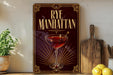 a decorative sign with the text "RYE MANHATTAN" and an illustration of a martini glass with a cherry on top, placed on a wooden surface next to a cutting board and some lemons.