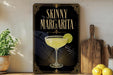 A framed poster of a skinny margarita cocktail is displayed on a wall, with a wooden cutting board and a potted plant to the side.
