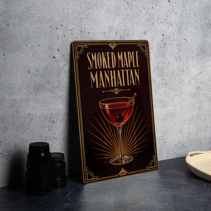 A framed poster of a smoked maple Manhattan cocktail is displayed on a gray wall, accompanied by a stack of black camera lenses and a white dish.