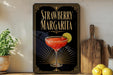 A framed poster of a strawberry margarita is displayed on a wall, with a wooden cutting board and a potted plant to the side.