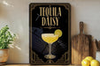 A decorative sign for "Tequila Daisy" cocktail, featuring a glass of tequila with a lime slice, placed on a wooden surface next to a cutting board and some lemons.