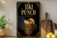 A framed poster of a tiki drink with a tiki mask and tropical fruits, displayed on a shelf next to a wooden cutting board and a potted plant.