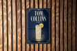 A sign for "Tom Collins" cocktail is displayed on a wooden wall.