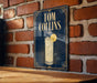 A sign for a cocktail named "Tom Collins" is displayed on a brick wall, featuring an illustration of a cocktail glass with a lemon slice.