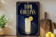 A framed advertisement for Tom Collins cocktails, featuring a glass of the drink and a lemon slice, is displayed on a wall next to a wooden cutting board and a potted plant.