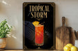 A decorative sign with the text "TROPICAL STORM" and an illustration of a tropical drink, placed on a wooden surface next to a potted plant and a cutting board.