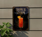 A sign for a tropical storm cocktail is displayed on a wall, with a plant visible to the left.