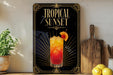 A framed poster of a tropical sunset cocktail is displayed on a wall, with a wooden cutting board and a potted plant to the side.
