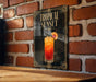 A framed poster of a tropical sunset cocktail is displayed on a brick wall.
