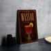 A framed illustration of a cocktail with a lemon slice, accompanied by the text "WASSAIL" on a dark background with a decorative border.