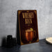 A metallic sign with the text "Whiskey Buck" and an illustration of a copper mug with a lime slice, placed on a gray countertop next to some black containers.