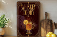 A decorative sign for whiskey tody, featuring a glass of whiskey with a lemon slice, is displayed on a shelf next to a wooden cutting board and a potted plant.
