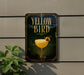 A sign for a cocktail called "Yellow Bird" is displayed on a wall, featuring an illustration of a cocktail glass with a garnish.