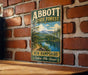 A vintage metal sign for Abbott State Forest in New Hampshire, featuring a scenic landscape with a lake and mountains.