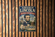 A vintage-style metal sign featuring the portrait of Abraham Lincoln, with the text "Abraham Lincoln National Heritage Area" and "Illinois" below the image.