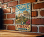 A vintage metal sign displaying the name "ACROPOLIS of Athens" and the word "GREECE" is mounted on a brick wall.