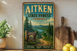 a vintage-style metal sign for Aitken State Forest in Vermont, with a scenic mountain landscape and a path leading through the forest.