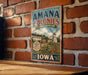 A vintage-style sign for "Amana Colonies Heritage Landscape" is displayed on a brick wall, featuring a windmill and a rural scene.
