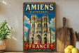 A vintage-style metal sign with the text "Amiens Cathedral" and "France" on it, featuring an illustration of the Amiens Cathedral. The sign is displayed on a shelf next to a wooden cutting board and some potted plants.