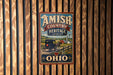 A vintage-style metal sign with the text "AMISH COUNTRY HERITAGE LANDSCAPE" and an illustration of a horse-drawn carriage, set against a wooden background.