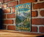 A vintage metal sign with the text "ANGELES NATIONAL FOREST" and "EXPLORE NATURE'S MAJESTY CALIFORNIA" is displayed on a brick wall.
