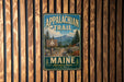 A vintage metal sign for the Appalachian Trail in Maine, featuring a scenic mountain landscape and a wooden signpost.