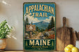 a vintage-style metal sign that reads "Appalachian Trail Maine Adventure Awaits", with a scenic mountain landscape in the background.