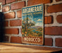 A vintage-style metal sign for the Arganie Biosphere Reserve in Morocco, featuring a scenic landscape with a tree, mountains, and a river.
