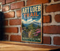 A vintage-style metal sign with the text "ART LOEB TRAIL NORTH CAROLINA" and a scenic mountain landscape is displayed on a brick wall.