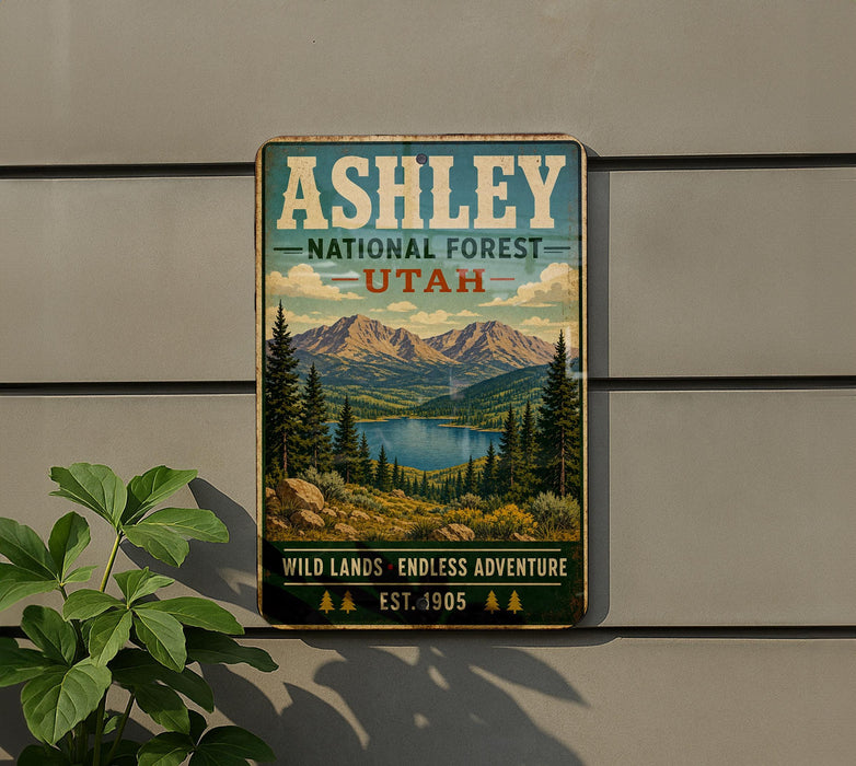 A vintage-style metal sign for the Ashley National Forest in Utah, featuring a scenic landscape with mountains, pine trees, and a lake.