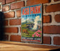A vintage-style sign for ASO Park, Japan, featuring a cow grazing in a mountainous landscape with a steam vent in the background.