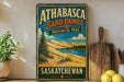 A vintage-style sign for Athabasca Sand Dunes Provincial Park in Saskatchewan, Canada, featuring a scenic landscape with sand dunes, trees, and a lake.