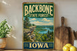 A vintage-style sign for the Backbone State Forest in Iowa, featuring a scenic landscape with a river and mountains.