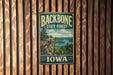 A vintage metal sign for the Backbone State Forest in Iowa, featuring a scenic landscape with a lake and mountains.