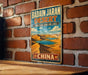 A vintage metal sign for Badain Jaran Desert, featuring a desert landscape with sand dunes and a lake, is displayed on a brick wall.