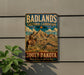 A vintage-style metal sign advertising Badlands Cultural Landscape South Dakota, featuring a cowboy on horseback and a scenic mountain landscape.