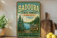 A vintage-style metal sign for Badoura State Forest in Minnesota, featuring a scenic illustration of pine forests, clear lakes, and a lake surrounded by mountains.