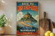 A vintage-style metal sign advertising the town of Mont Saint-Michel in France, featuring a castle and beach landscape.