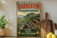 A vintage-style metal sign advertising Barberton, a mountain range in South Africa, with a scenic mountain landscape and text inviting viewers to explore the region's oldest mountains.